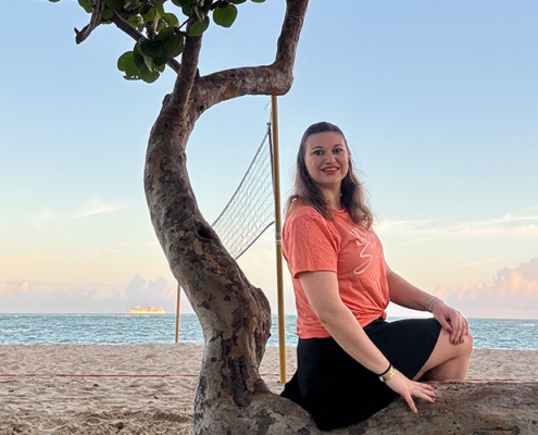 Anastasia Taran sitzt am Strand auf einem Baum, dahinter ein Volleyballfeld