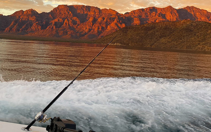 Hochseefischen in Bahía de Loreto, eine Angel ist am Boot befestigt und im Hintergrund sieht man Berge, die von der aufgehenden Sonne angestrahlt werden